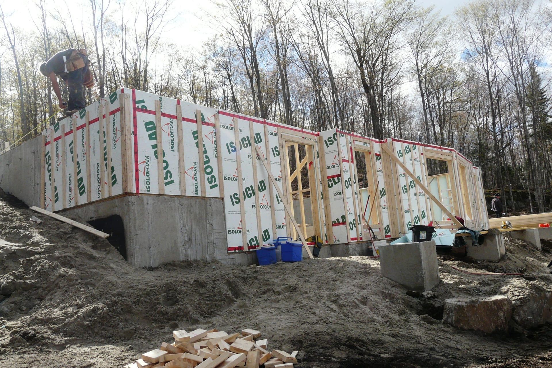 Une personne se tient debout sur le mur à ossature d'un bâtiment en construction, constitué de panneaux isolants reposant sur une base en béton.