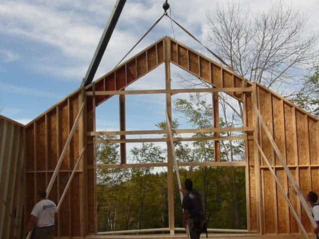 Des ouvriers utilisent une grue pour soulever une grande structure murale en bois pour une maison en construction à l'extérieur.