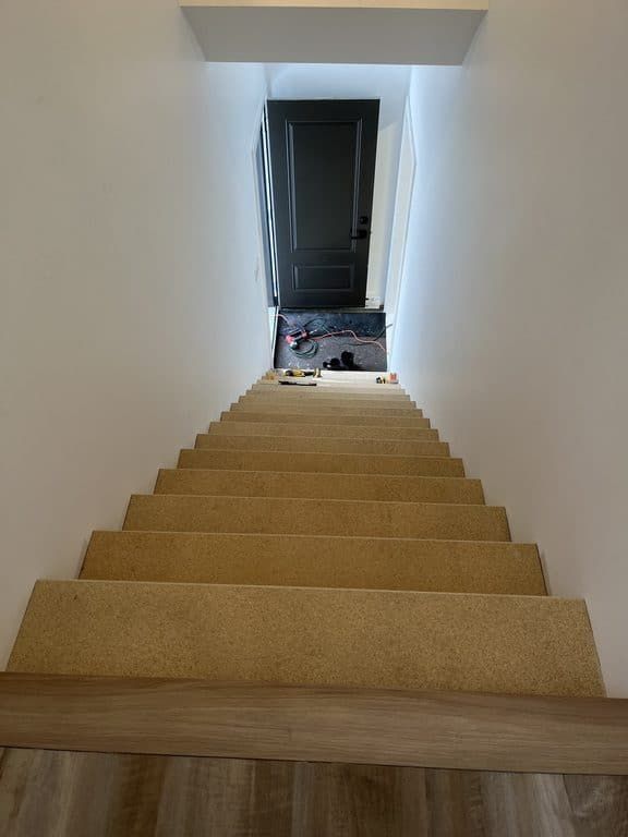 Un escalier droit recouvert de moquette claire, vu du haut en direction d'une porte sombre en bas.