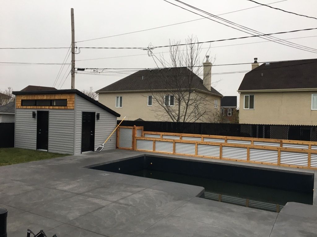 Dans le jardin, un abri de jardin gris jouxte une piscine rectangulaire entourée d'une clôture en tôle ondulée, le tout sur une terrasse en béton.