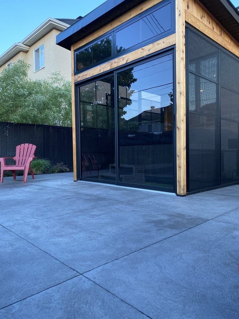 Une structure extérieure moderne en bois, dotée de grands panneaux de verre et d'une paroi vitrée, se dresse sur une terrasse pavée avec une chaise rose.