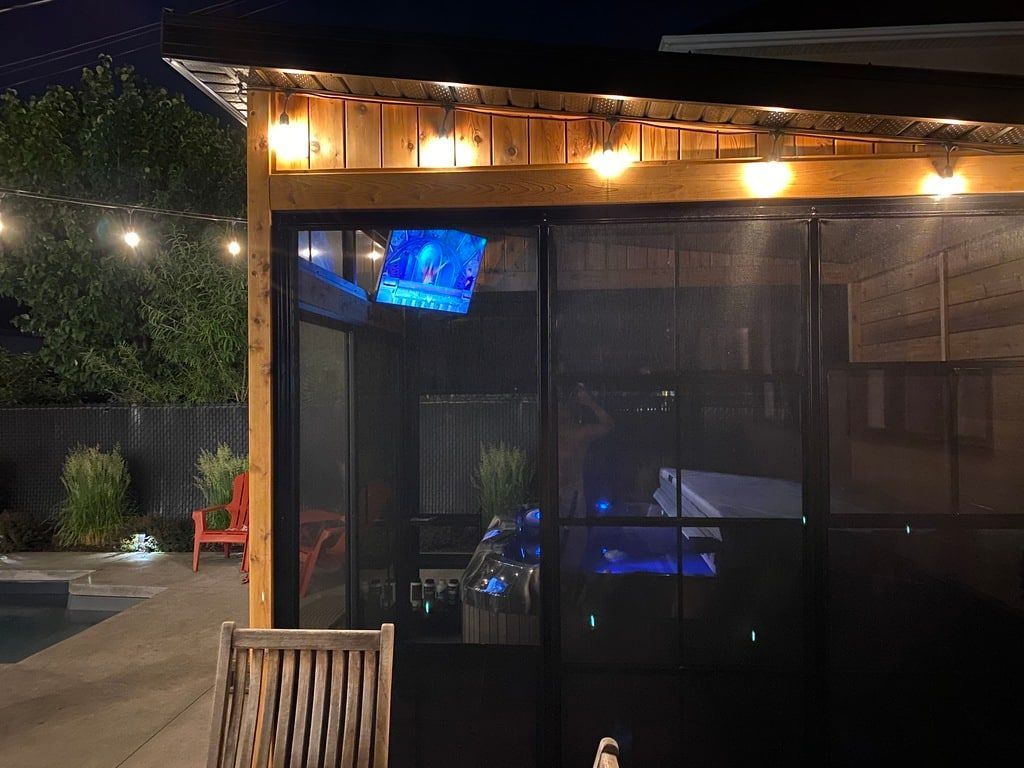 Vue nocturne d'une terrasse en bois illuminée par des guirlandes lumineuses, abritant un jacuzzi illuminé et une télévision murale.