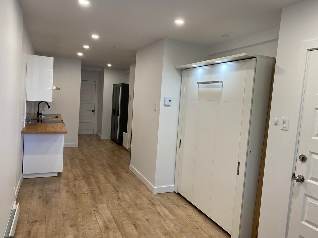 Un petit studio moderne avec un lit escamotable blanc, un parquet et un coin cuisine compact.