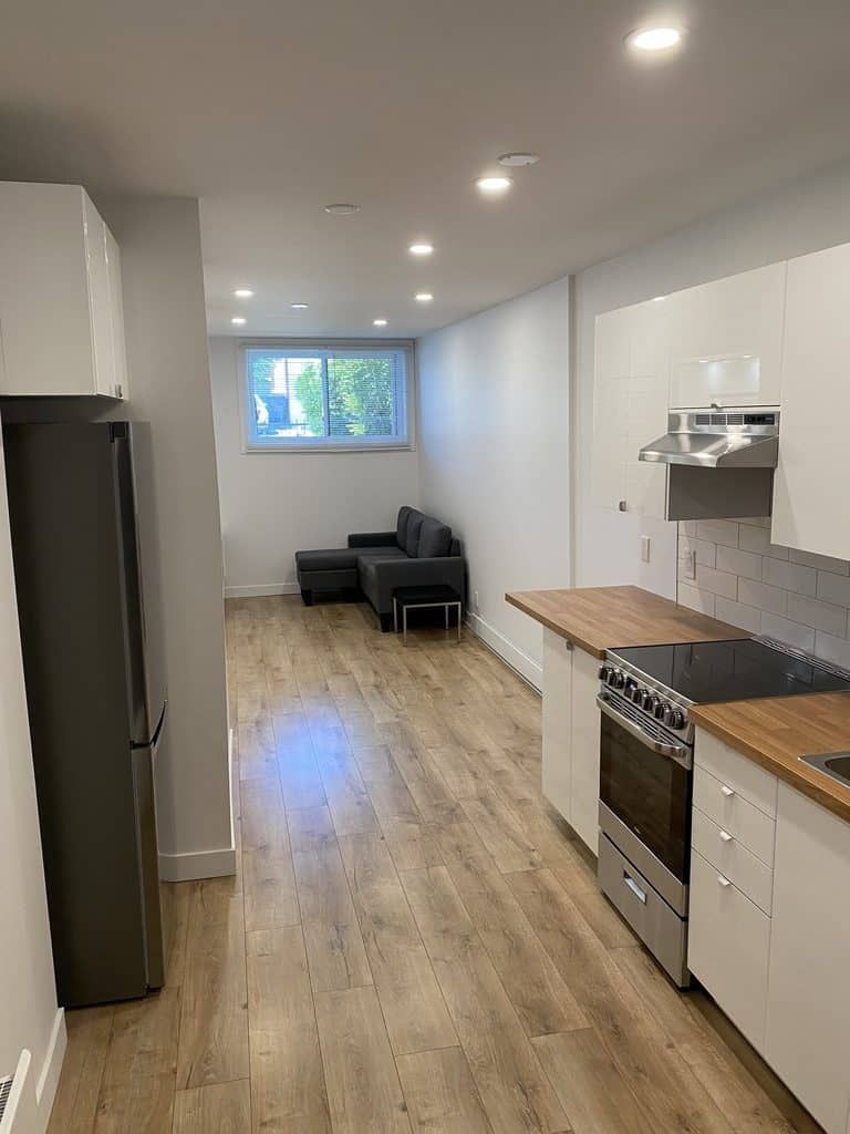 Une cuisine et un séjour modernes et étroits, à aire ouverte, avec des armoires blanches, des comptoirs en bois et un canapé gris.