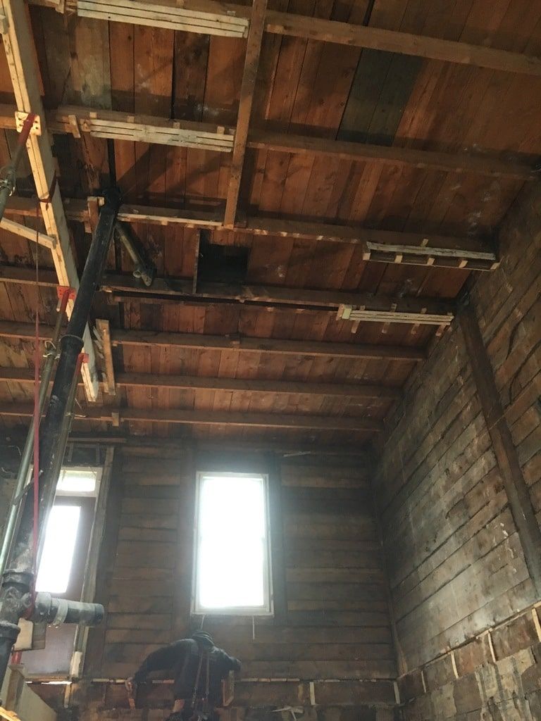 Vue intérieure d'une pièce en construction avec des solives de plafond en bois apparentes, des murs en bois foncé et deux fenêtres.