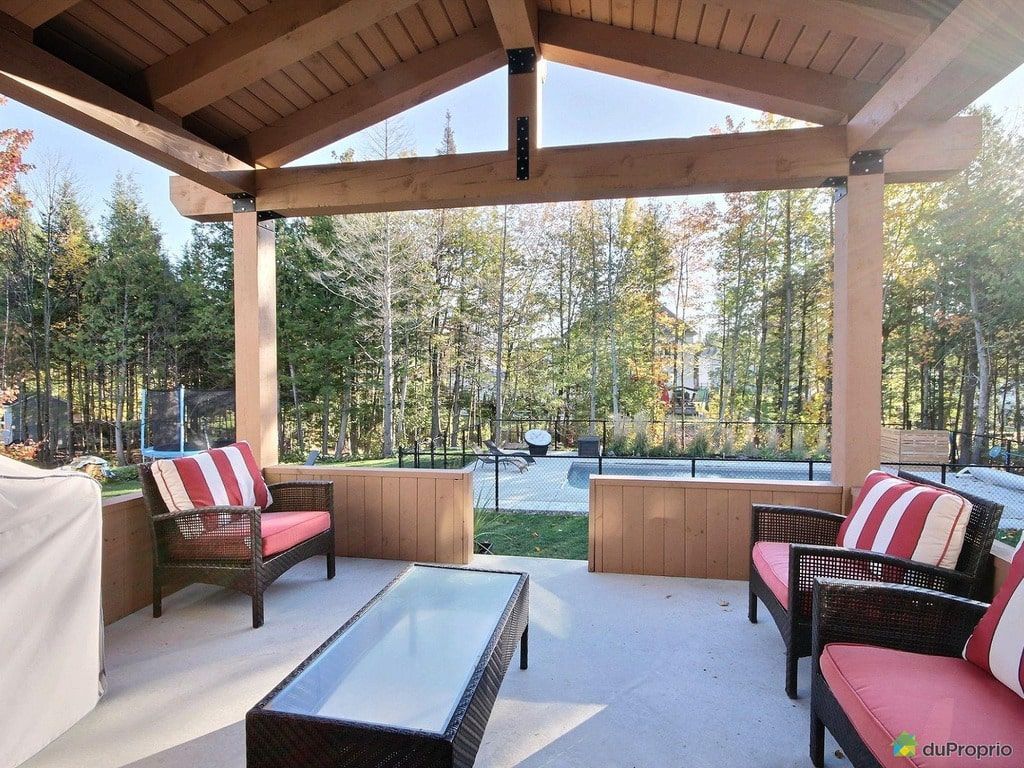 Terrasse avec une structure en bois couverte, des sièges avec des coussins rouges et blancs, une table en verre et une vue sur une piscine et des arbres.