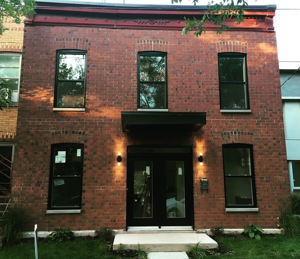 Un bâtiment de deux étages en briques rouges avec des fenêtres à encadrement noir, une entrée principale centrale et un auvent de porte noir moderne.