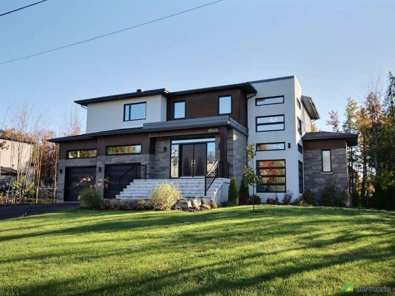 Une maison moderne de deux étages, avec un enduit blanc, des éléments en bois et un revêtement en pierre, se dresse sur une pelouse avec un escalier d'entrée en béton.