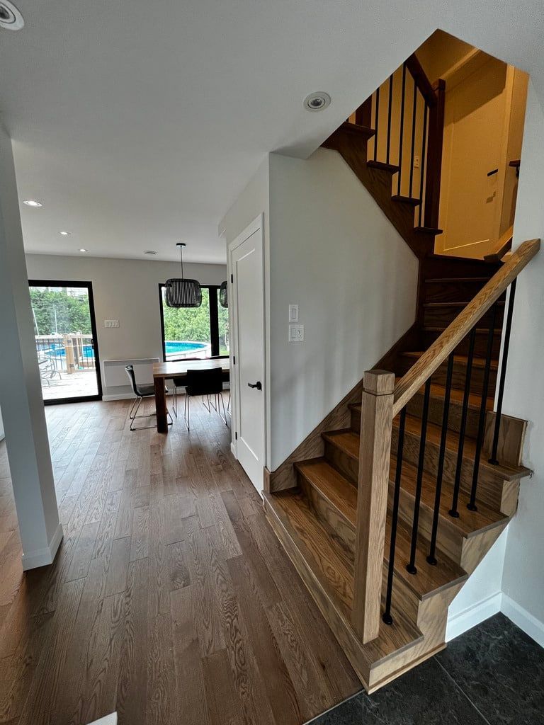 Vue d'une entrée de maison avec parquet, un escalier aux marches en bois et à la rampe noire, et un coin repas.