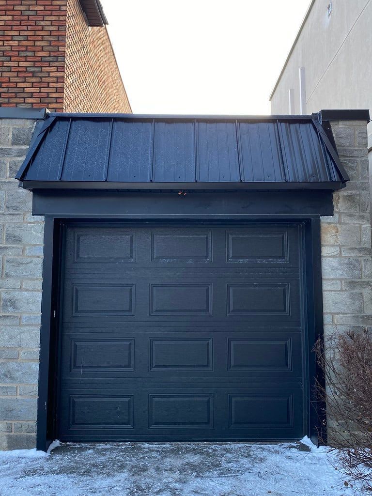 Une porte de garage noire se trouve sous un auvent à toit métallique noir, flanquée de murs en pierre.