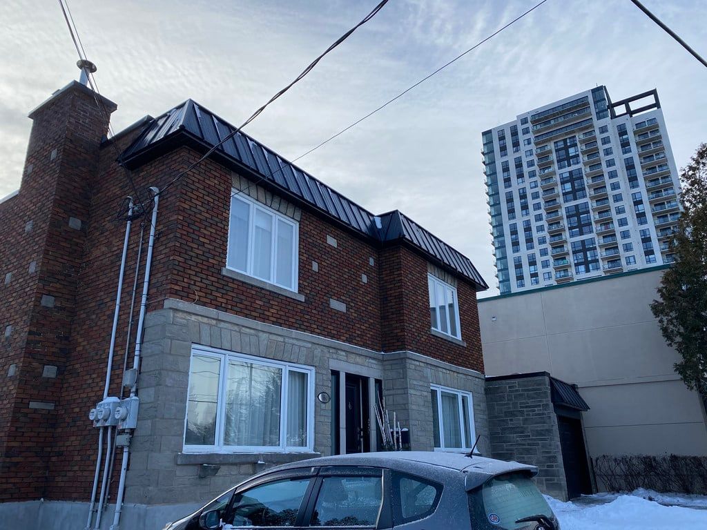 Sous un ciel nuageux, une maison en briques au toit métallique se dresse à côté d'un grand immeuble d'appartements moderne.