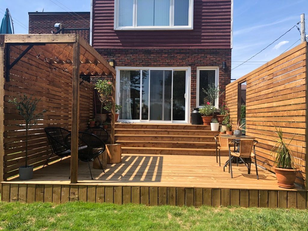 Une terrasse en bois à plusieurs niveaux, aménagée avec des lattes pour préserver l'intimité, du mobilier de jardin et des plantes en pot, attenante à une maison en briques.