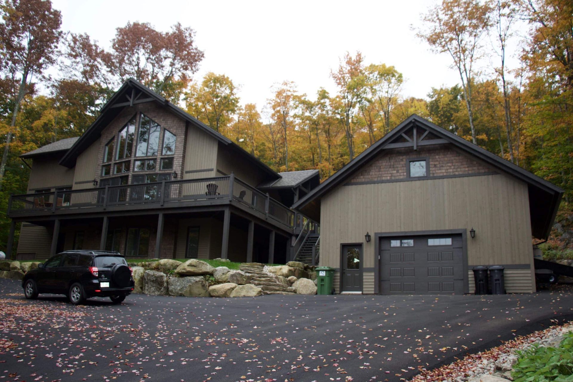 Une maison brune de deux étages avec une terrasse et un garage attenant, se détachant sur un fond d'arbres d'automne le long d'une allée pavée.