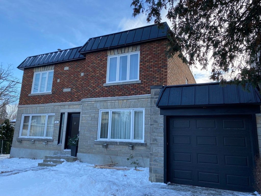Une maison à deux étages avec des murs supérieurs en briques brunes, des murs inférieurs en pierre grise, un toit en métal sombre et un garage sombre attenant.