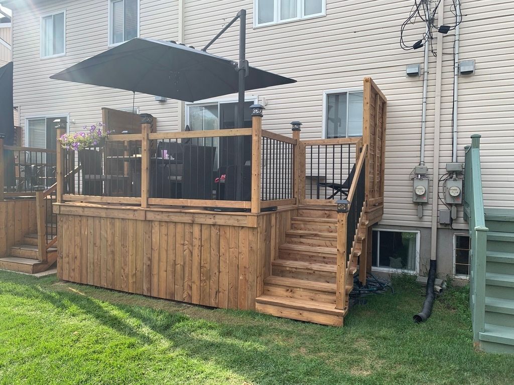 Une terrasse en bois avec des rambardes en métal noir et un parasol ouvert, reliée à une maison beige par un petit escalier.