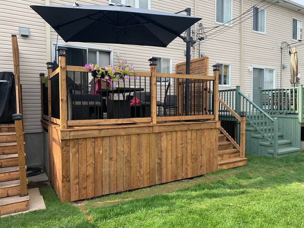 Une terrasse en bois avec des balustrades noires, un grand parasol bleu marine et du mobilier de jardin dans une cour arrière résidentielle.