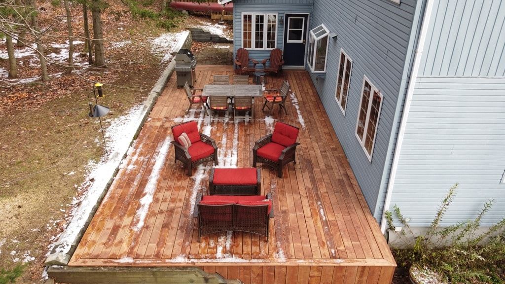 Vue en plongée d'une terrasse en bois donnant sur un jardin, avec un ensemble de salle à manger et deux chaises longues rouges, près d'une maison aux murs bleus.