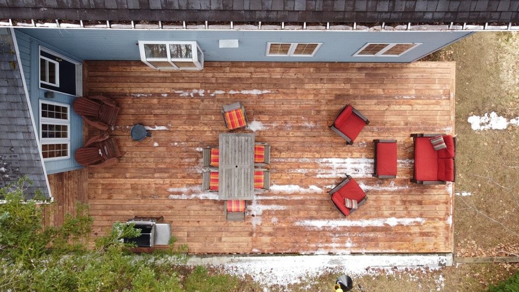 Vue aérienne d'une terrasse rectangulaire en bois avec mobilier de jardin, chaises et barbecue, attenante à une maison bleue.