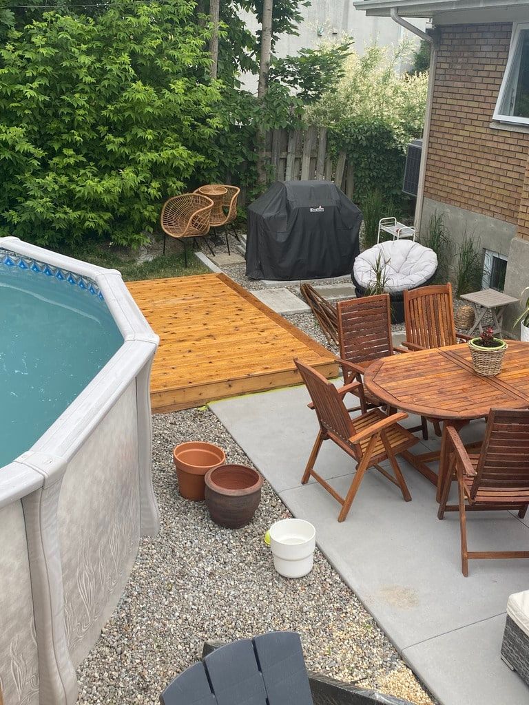 Une scène de patio dans une cour arrière avec une piscine hors sol, une terrasse en bois, des meubles de patio et des plantes en pot sur une surface gravillonnée.