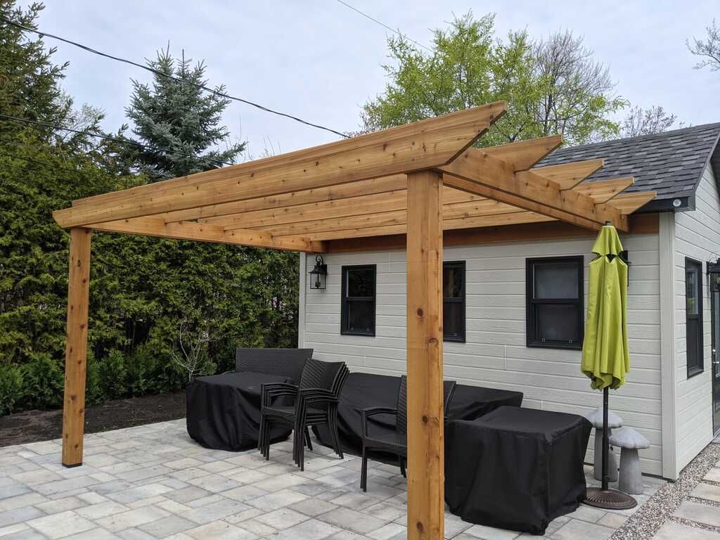 Une pergola en bois surplombe une terrasse en pierre avec des meubles de jardin couverts, à côté d'un bâtiment de stockage de couleur claire.