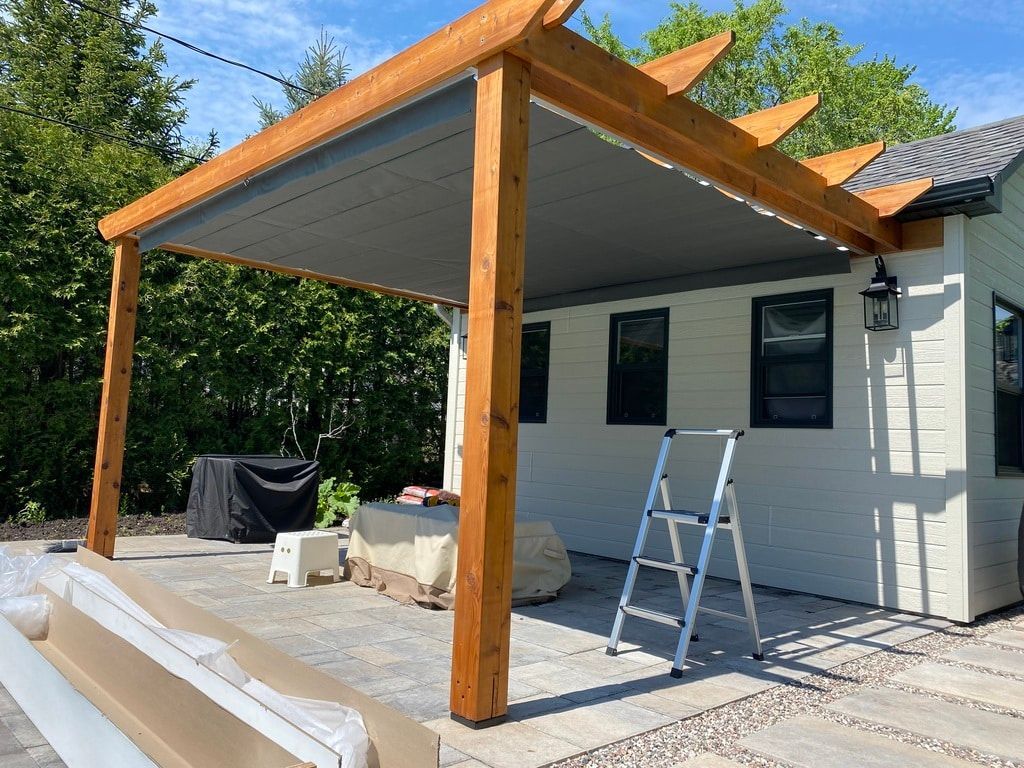 Une pergola en bois avec un toit rétractable en tissu gris surplombe une terrasse en pierre à côté d'une maison blanche avec une échelle métallique.