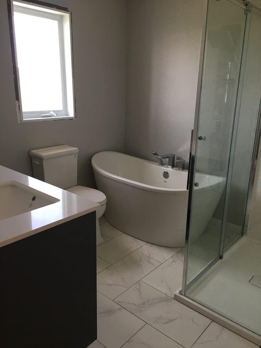 Une salle de bains moderne avec une baignoire ovale blanche sur pieds, des toilettes, une cabine de douche avec portes en verre et un sol imitation marbre.