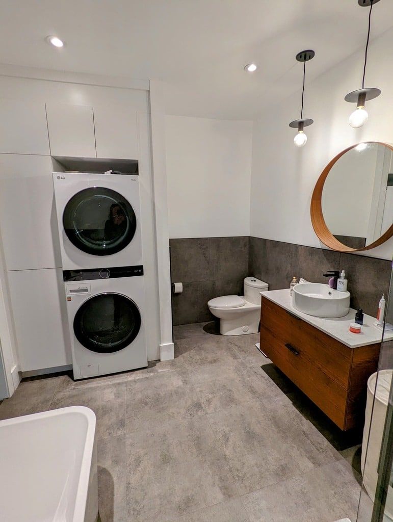 Une salle de bains moderne comprenant un lave-linge et un sèche-linge superposés, un meuble-lavabo en bois avec un lavabo vasque blanc et un miroir mural circulaire.