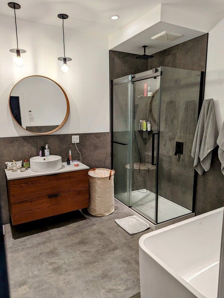 Salle de bains moderne avec meuble-lavabo en bois, miroir rond, douche vitrée, panier à linge clair et baignoire blanche.