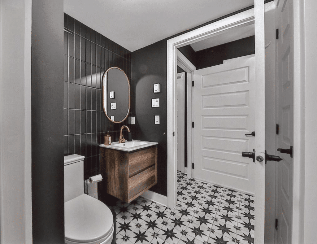Des toilettes avec des murs carrelés de carreaux de métro noirs, un meuble-lavabo en bois, un miroir rond et un carrelage au sol à motifs noirs et blancs.