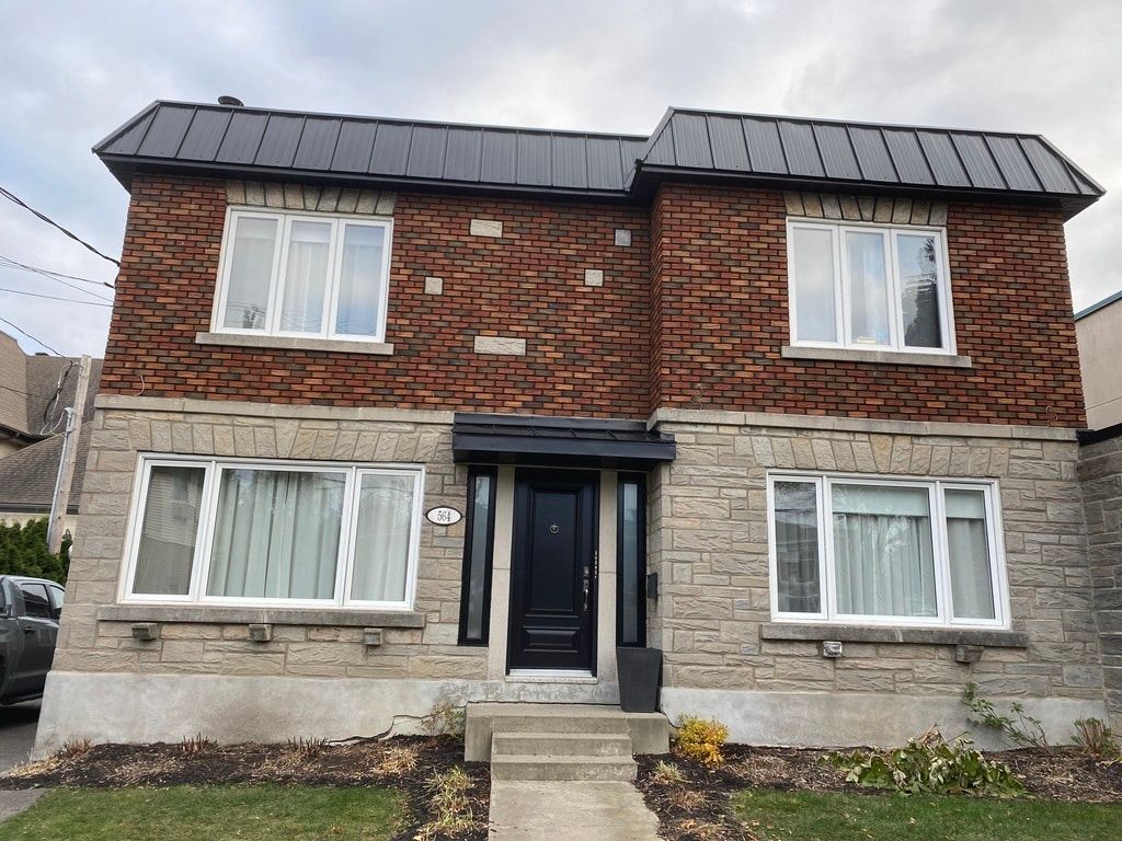 Maison de deux étages en briques et en pierres, avec une porte d'entrée noire et un toit en métal, sous un ciel nuageux.