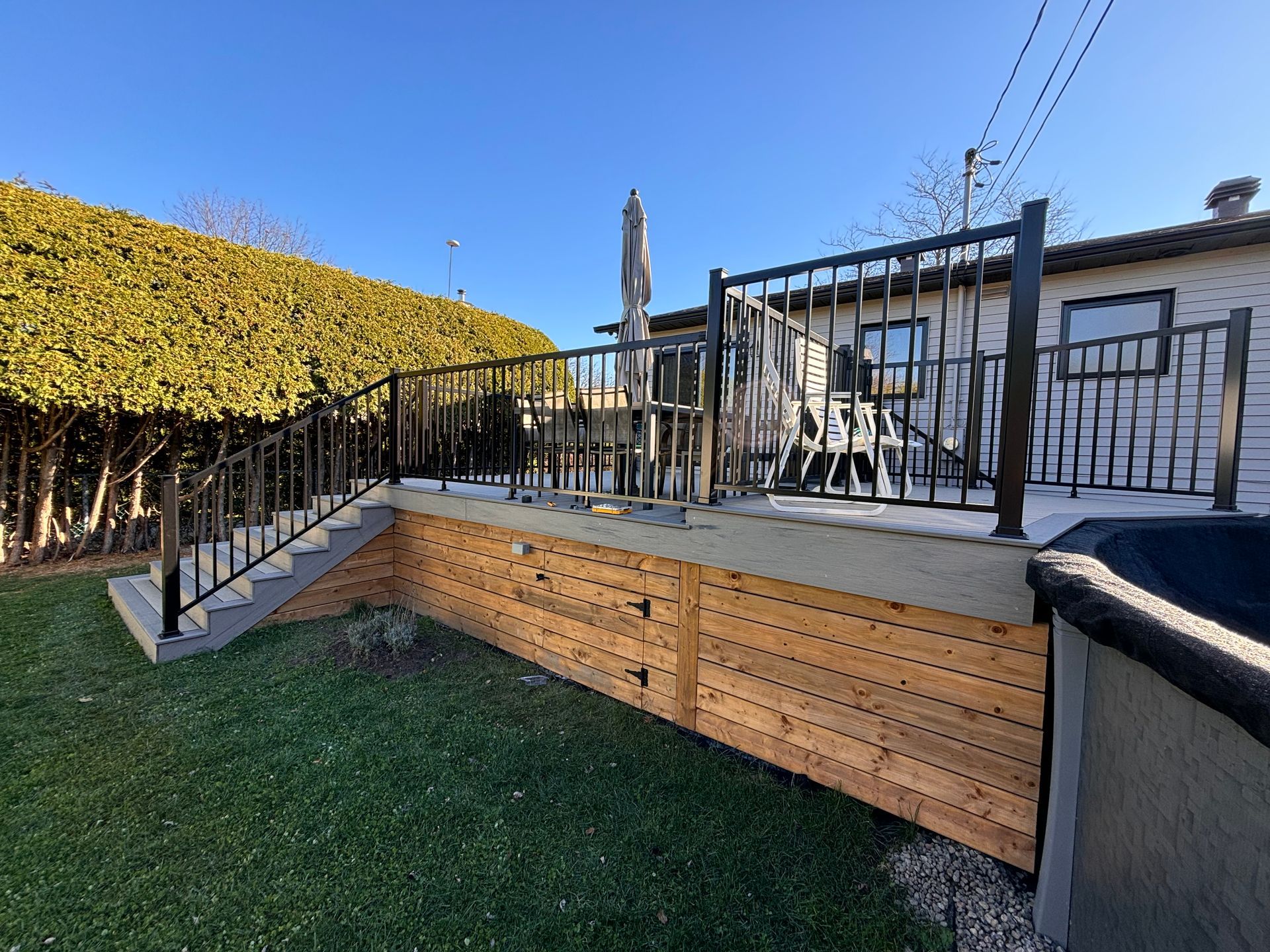 Une terrasse extérieure surélevée en bois avec des rambardes et des escaliers en métal noir, adossée à une maison et à une grande haie verte.