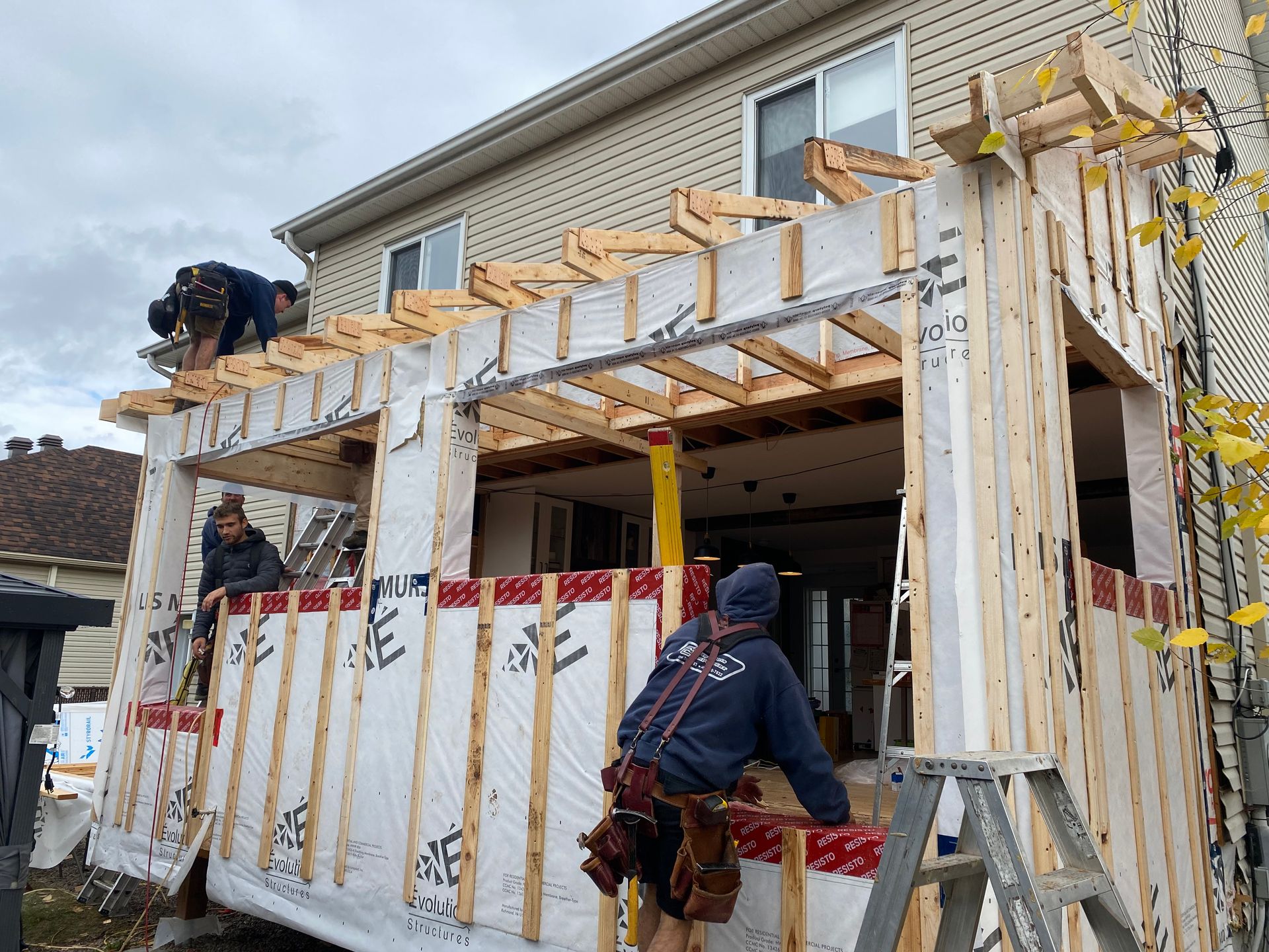 Deux ouvriers du bâtiment construisent une extension à ossature bois à l'arrière d'une maison beige à deux étages.
