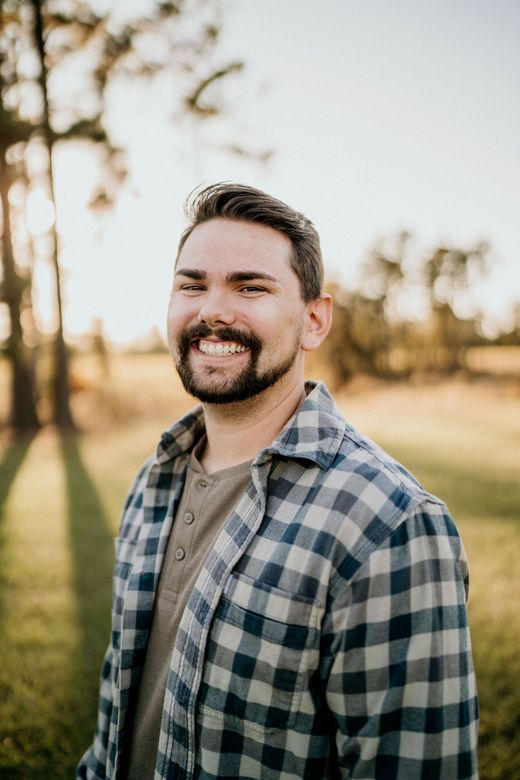 A man with a beard is wearing a plaid shirt and smiling in a field.