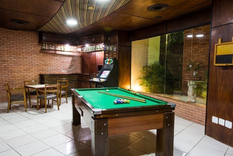 Sala de jogos com painéis de madeira, mesa de bilhar, cadeiras e máquina de jogos arcade sob iluminação aconchegante.