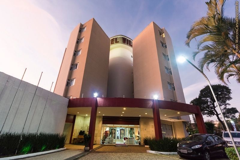 Entrada de um prédio moderno ao entardecer, com altas torres curvas, janelas iluminadas e palmeiras em frente.