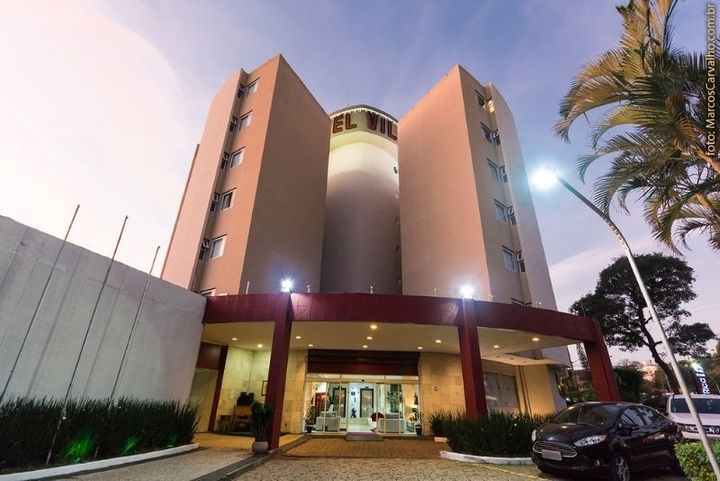 Entrada de um prédio moderno ao entardecer, com altas torres curvas, janelas iluminadas e palmeiras em frente.