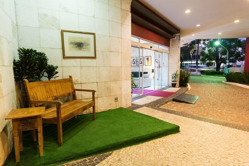 Entrada coberta com banco de madeira, tapete verde e passarela aberta à noite.