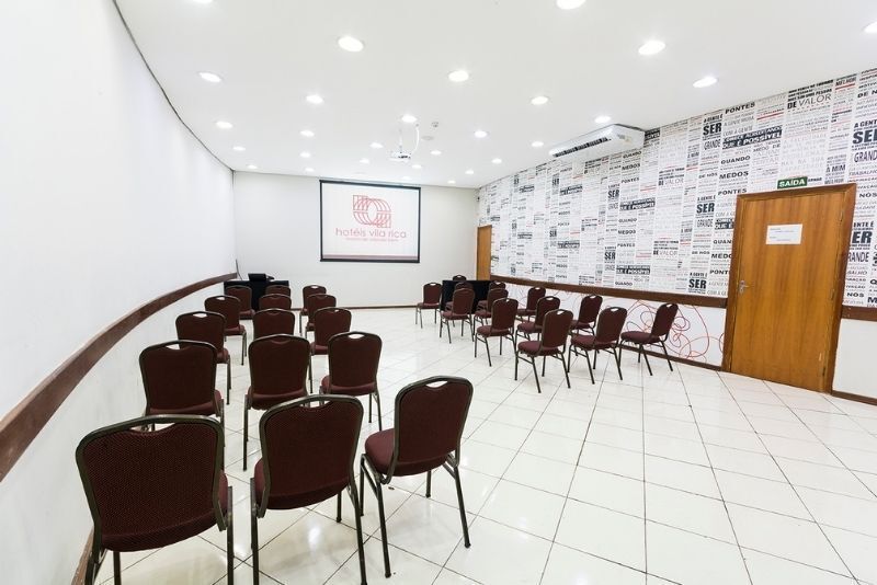 Sala de reuniões vazia com fileiras de cadeiras marrons, piso de azulejos brancos e uma porta de madeira à direita.