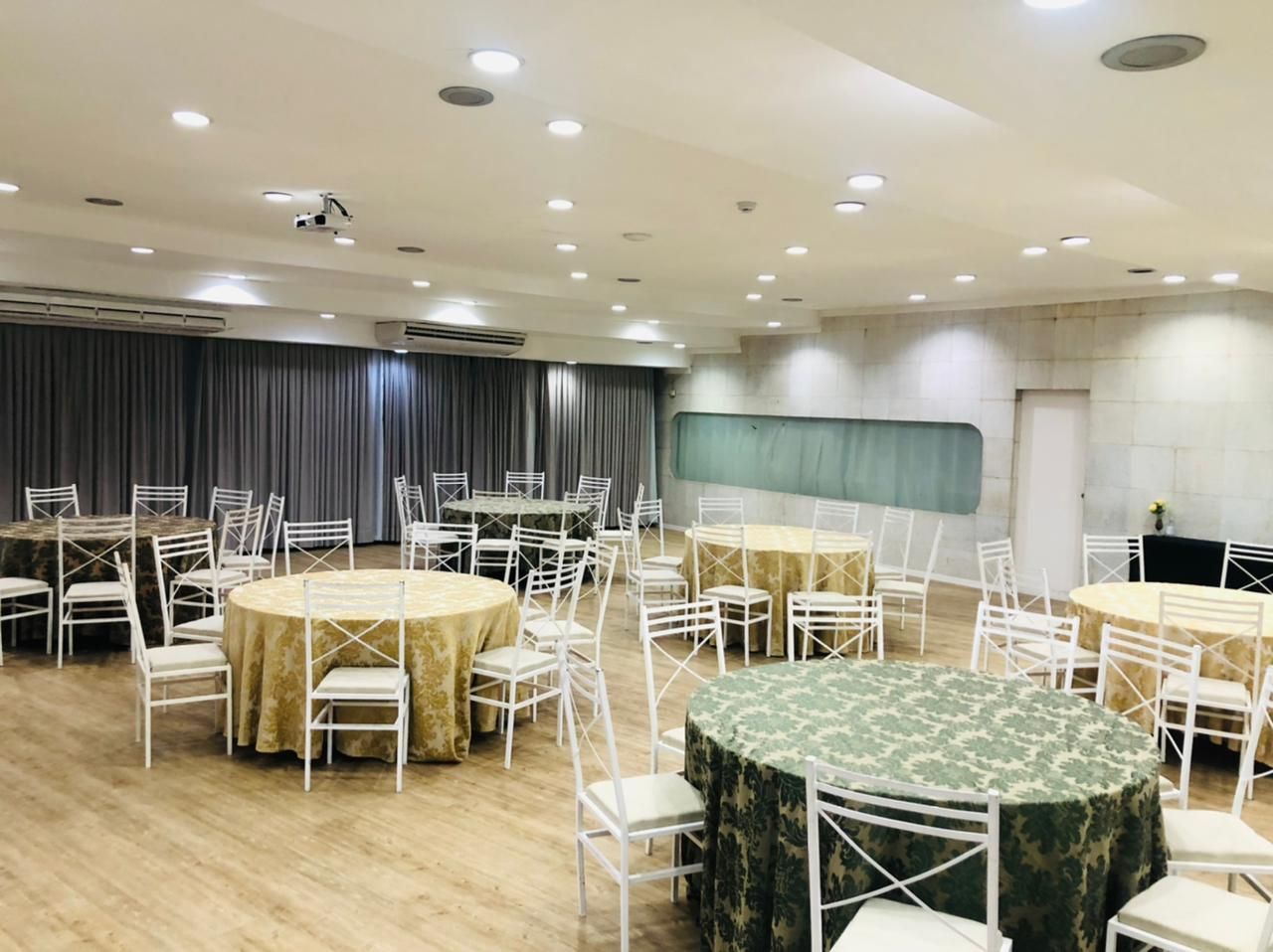 Salão de banquetes vazio com mesas redondas cobertas com toalhas de mesa douradas e verdes e cadeiras brancas sob luzes de teto.