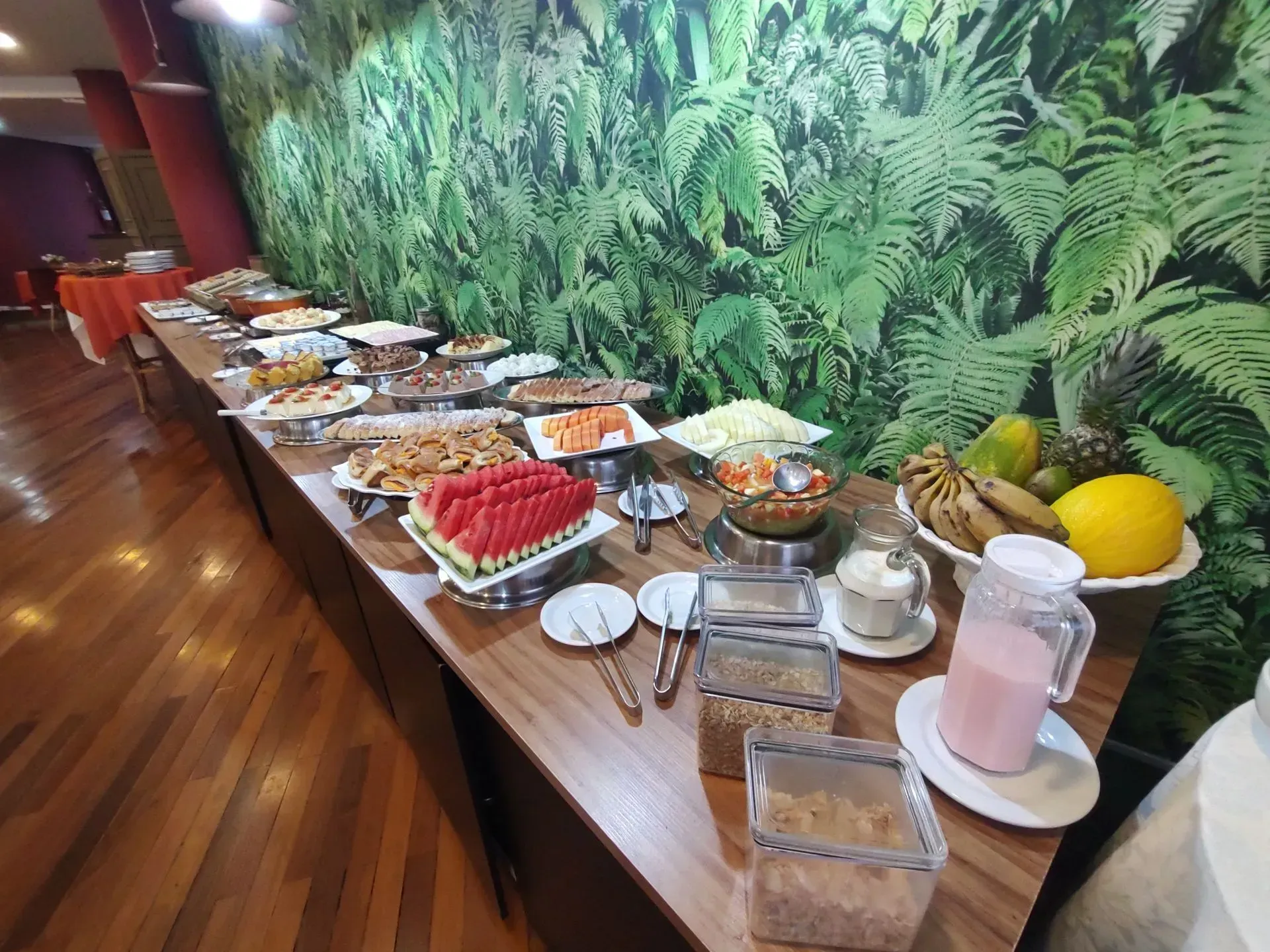 Mesa de buffet com pratos variados, frutas e bebidas contra uma parede verdejante em uma sala de jantar.