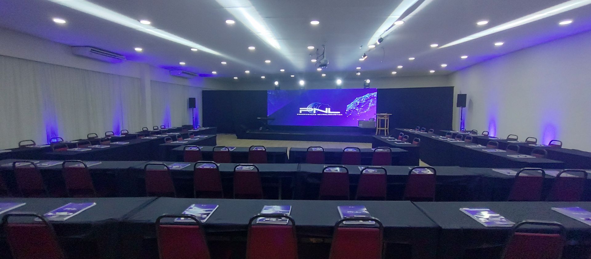 Amplo salão de conferências com fileiras de mesas voltadas para um palco iluminado e um telão.