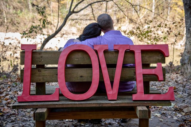 Duas pessoas sentadas em um banco atrás de grandes letras vermelhas que formam a palavra AMOR em um ambiente arborizado ao ar livre.
