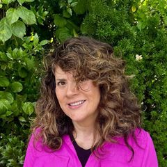 Woman with curly brown hair, smiling, wearing a pink jacket, in front of green foliage.