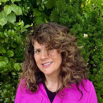 Woman with curly brown hair, smiling, wearing a pink jacket, in front of green foliage.