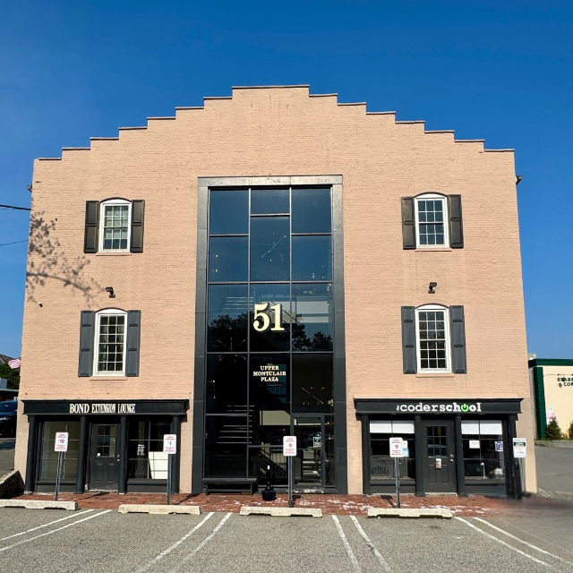 A tan, three-story brick building with a central glass entryway numbered 51, featuring ground-floor shops and parking.
