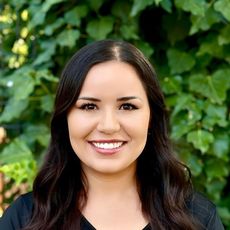Woman with curly brown hair, smiling, wearing a pink jacket, in front of green foliage.