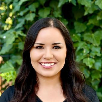 Woman with curly brown hair, smiling, wearing a pink jacket, in front of green foliage.
