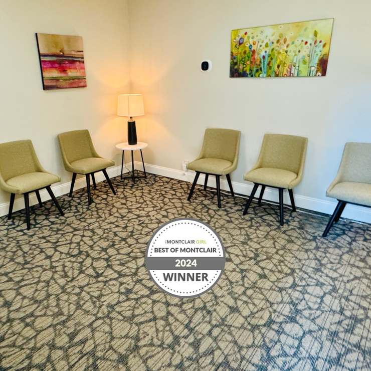 A waiting room with five chairs, carpet flooring, artwork, and a side table