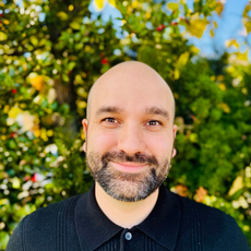 A person with a shaved head and facial hair, smiling gently while wearing a black collared shirt in front of greenery.