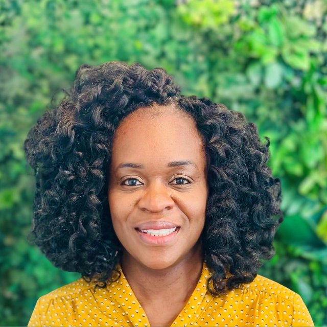 Smiling woman with curly hair in a yellow polka-dot top against a green leafy background